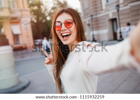 Selfie-portrait of pretty girl with long brunette hair at street background. wears beige dress, red sunglasses, make up. Smiling and positive.young woman in glasses making selfie , showing two fingers