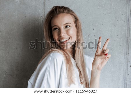 Picture of amazing beautiful young woman indoors over grey wall looking aside showing peace gesture.