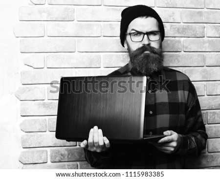 Bearded man, long beard. Brutal caucasian serious unshaven hipster holding laptop in red black checkered shirt with hat and glasses on beige brick wall studio background