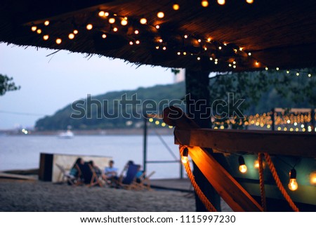Summer bar on the river bank, decorated with light bulbs and lanterns. People relax in the sun loungers. Party, summer evening.  beautiful illumination. city beach. Royalty-Free Stock Photo #1115997230