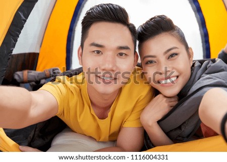 Portrait shot of adorable Asian couple taking selfie while lying in tent after busy day outdoors, first person view
