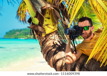 Male Paparazzi hides behind a tree on a tropical beach to take pictures of a hidden camera
