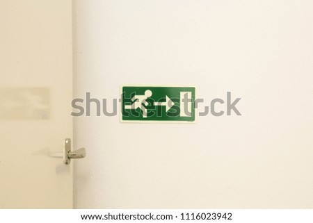 The emergency exit sign shows the direction of escape in case of danger. The emergency exit board hangs on the ceiling of the building. white wall