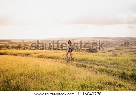 Girl with a bicycle posing on the hill in beautiful rural landscape at sunset. Young pretty female person with retro bike standing in a meadow on bright sunny afternoon in summer
