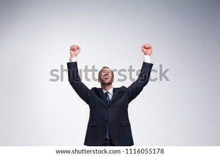 Studio portrait of young handsome businessman in black suit showing victory gesture isolated on light blue background
