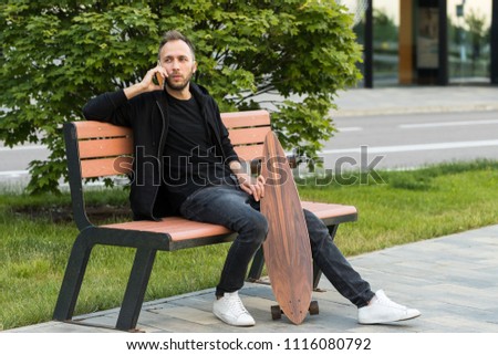 Attractive European man sitting with skateboard or longboard on the bench, using smart phone and relaxing.Leisure, healthy lifestyle, extreme sports concept.Stylish skateboarder with wooden longboard