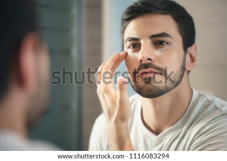 Young hispanic people and male beauty. Metrosexual man applying lotion for anti-aging treatment around eye Royalty-Free Stock Photo #1116083294