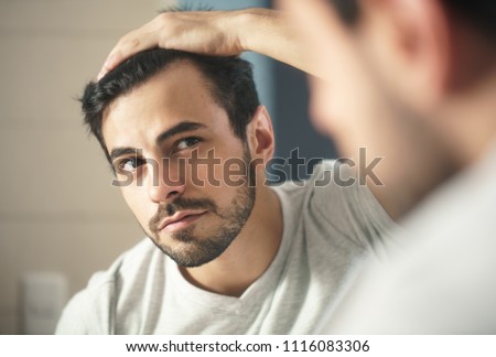 Latino person with beard grooming in bathroom at home. White metrosexual man worried for hair loss and looking at mirror his receding hairline. Royalty-Free Stock Photo #1116083306