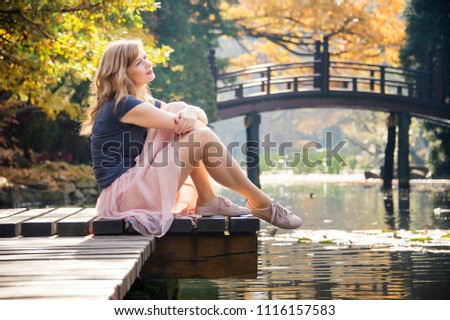 dreaming young blond woman relaxed on a pier near lake in autumn city park in a warm fall sunny day