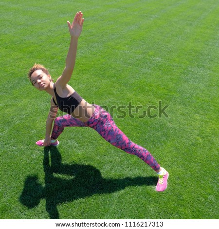 Modern style of life. A young African-American girl in a black T-shirt, pink pants and sneakers doing sports exercises on green grass and raising her hands up. Square picture.