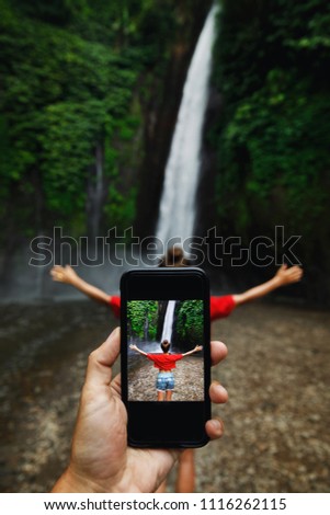 Male hand with a smartphone. Man taking picture of her girlfriend beside beautiful waterfall.