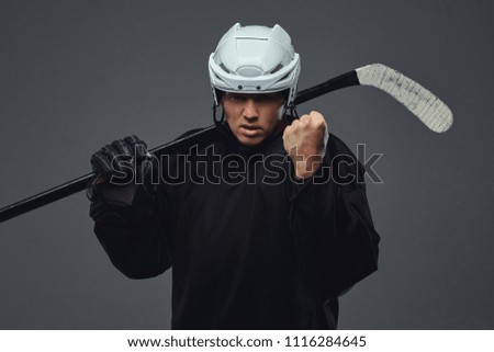 Angry professional hockey player in black sportswear standing with a hockey stick on a gray background.