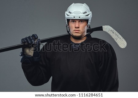 Hockey player wearing black protective gear and white helmet holds a hockey stick on a gray background.