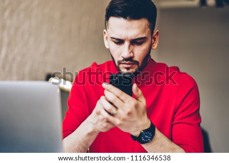 Concentrated male blogger installing app on smartphone using wireless internet connection during work at laptop computer.Pensive bearded young man reading news on website on mobile phone