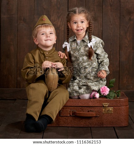 children boy are dressed as soldier in retro military uniforms and girl in pink dress sitting on old suitcase, dark wood background, retro style