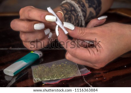 girl sitting at a table makes a narcotic cigarette, a lighter and a packet with a weed lie on a table near