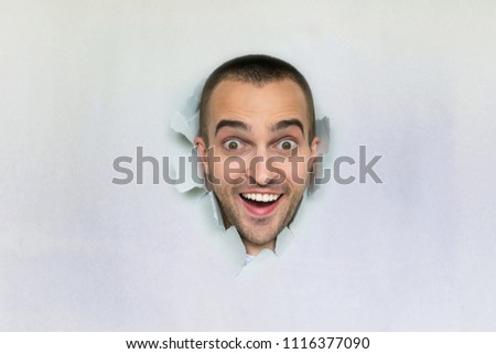 Handsome young guy making hole in paper, looking at the camera, smiles widely, contented, blue background with a copy of the space, for advertising