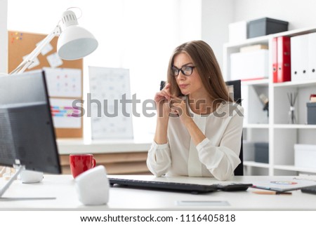 A young girl is sitting at the desk in the office, holding a pencil in her hand and looking at the monitor.