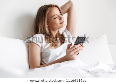 Photo of pretty young woman indoors at home using mobile phone chatting. Stretching lies in bed.