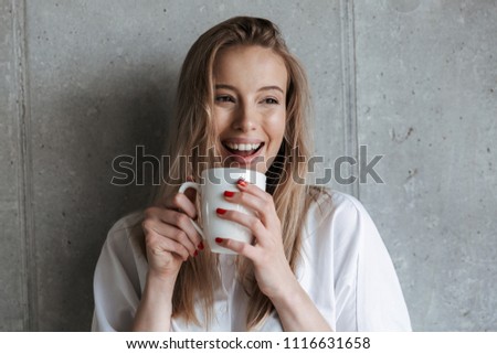 Picture of cute happy young woman indoors over grey wall drinking coffee holding cup.
