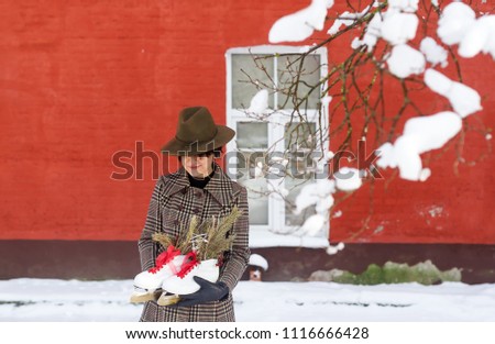 Portrait of a beautiful young brunette in big hat standing with skates in her hands against the background of the house from a red brick.