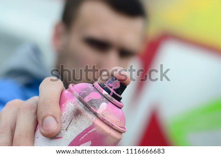 A young graffiti artist in a blue jacket is holding a can of paint in front of him against a background of colored graffiti drawing. Street art and vandalism concept