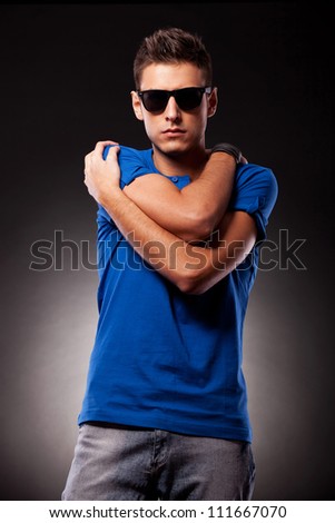 Waist-up picture of a young casual man with sunglasses holding his hands folded with elbows in front and palms on his shoulders, while facing the camera