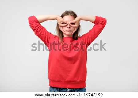 Pretty girl in red sweater holding fingers near eyes like glasses: mask like super hero or owl