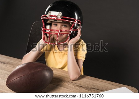 little smiling boy in protective helmet at table with american football ball on grey background 