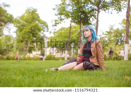 Beautiful Russian young woman sits on a lawn in a kurta and black clothes. Holds a plastic reusable glass with a cold drink.