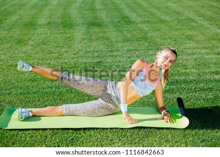 Blonde positive fitness woman doing legs rise on the green grass outdoors at stadium. Sport in open air and health care concept.