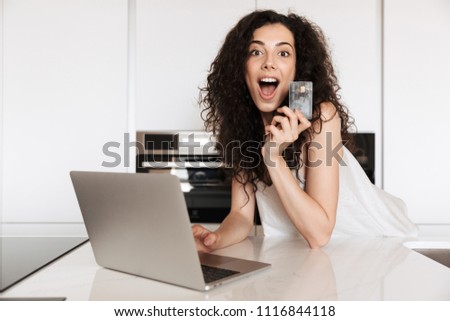 Picture of excited young woman 20s with curly brown hair wearing silk leisure clothing shouting in happiness and shopping online with laptop and credit card while leaning on kitchen table