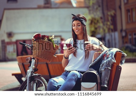 Smiling young woman texting on smart phone on the city street, on a sunny day, beside the bike with flower basket.