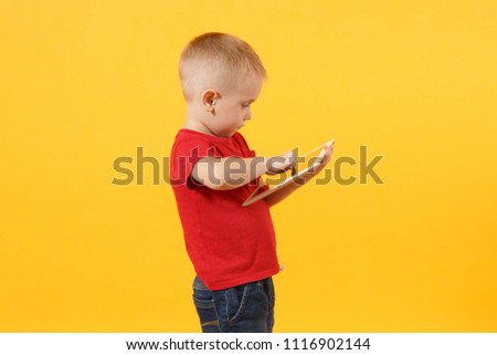 Little cute kid baby boy 3-4 years old in red t-shirt holding in hand tablet pc computer isolated on yellow background. Kids childhood lifestyle concept. Problem of children and gadgets. Copy space