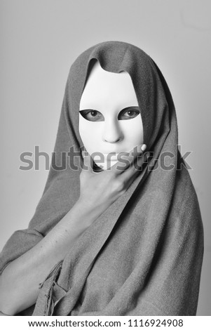 Black and white photo of a woman in a white mask and grey scarf, her baleful stare pirces ones soul