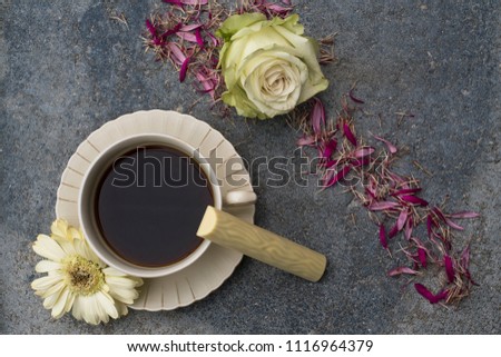 A cup with coffee stands in front of dark concrete background. Champagne colored flowers and floral petals decorate the picture. A candy made of white chocolate tempted to a coffee break.