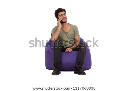 Handsome young man wearing a casual outfit, sitting on a bean bag, holding his phone talking to someone, isolated on white background