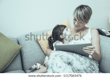 Modern mother showing educational film to daughter. Pretty girl looking at mom. They relaxing on sofa in living room. Positive family using technology. Leisure concept