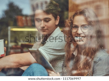 Couple of young students drinking coffee and using a digital tablet while sitting on a window sill at a college campus during a break.