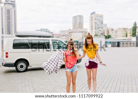 Slim girls with long legs looking down, standing on main city square with skyscrapers on background. Outdoor portrait of gorgeous young sisters walking together on the street with amazing view.
