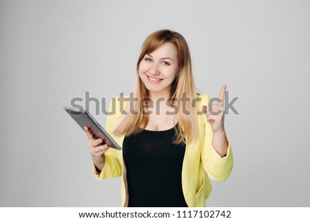 Portrait of creative blonde haired woman, wearing in yellow jacket and black blouse, holding tablet computer and pointing finger up against white studio background. Concept of business ideas.