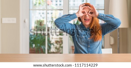 Redhead woman at home doing ok gesture like binoculars sticking tongue out, eyes looking through fingers. Crazy expression.