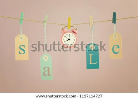 On a light pink background on the rope hang labels labeled SALE. In the center is a small pink alarm clock. Beautiful picture, concept, time for sales.