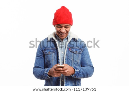 Cheerful black male model in denim jacket surfing new smartphone and looking happy isolated on white. 