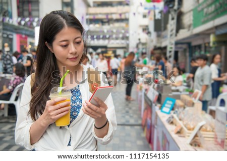 Asian woman holding a cup of mango juice and looking for the merchandise beside the road. She use her cell phone to take the picture of them.