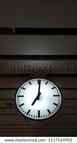 Hanging illuminated clock under dark ceiling of subway station at 7:01.