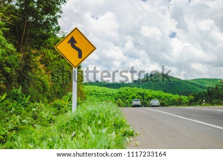 Warning reverse curve left traffic sign with vehicles on road. Landscape view of roadside at countryside in Thailand.