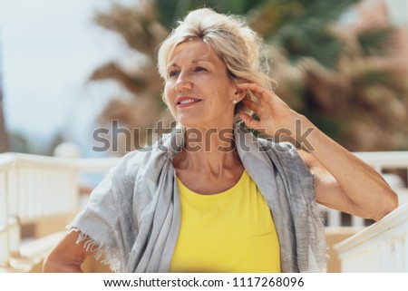 Elegant older blond woman on an outdoor patio posing with her hand to her hair and a friendly smile looking to the side