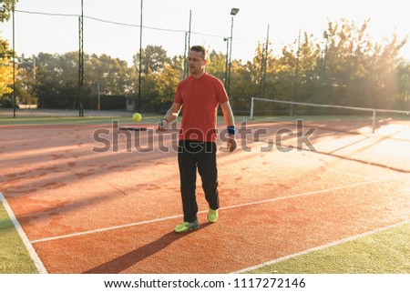 Concept health and sport. Athletic man warming up hitting the ball with racket, wearing sport clothes on tennis court background outdoors.