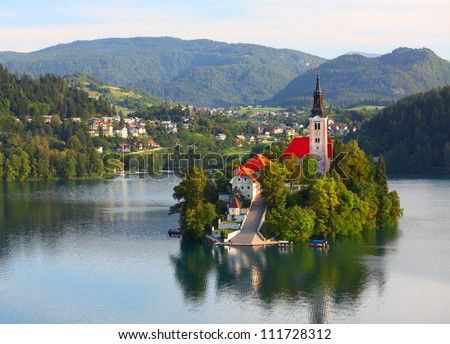 Catholic church situated on an island on Bled lake with mountains and villages on the background Royalty-Free Stock Photo #111728312